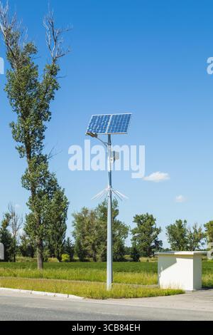 Modern solar panel streetlight stands beside a small rural bus shelter, surrounded by greenery and clear blue sky. Stock Photo