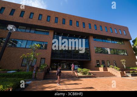 Exterior view of Kyoto University Library building with large glass windows, surrounded by greenery and illuminated by bright daylight, while visitors Stock Photo