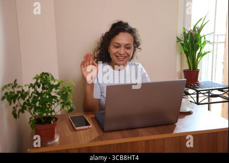 Smiling woman engaging in a virtual meeting on a laptop in a cozy, plant-adorned home setting, showcasing remote work and communication. Brings a sens Stock Photo