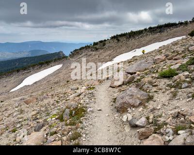 A lone hiker explores Mount Hood Timberline trail, surrounded by rocky terrain, patches of snow, and expansive vistas under a dramatic cloudy sky, evo Stock Photo