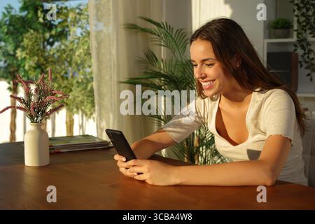 Happy woman using mobile phone on wooden table at sunset in the living room at home Stock Photo