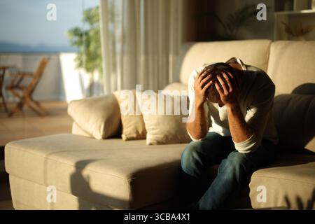 Sad and desperate man sitting on a couch at home at sunset Stock Photo