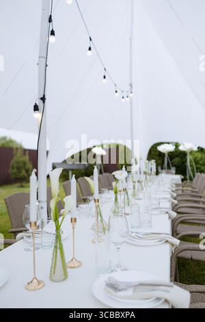 Outdoor wedding table with white linens, gold candle holders, and calla lily vases under a white tent with string lights Stock Photo