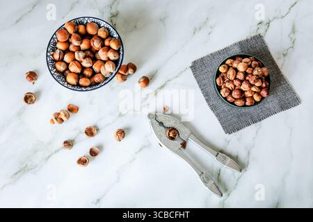 Rustic flat-lay of whole and shelled hazelnuts with vintage metal nutcracker on white marble countertop, shot in natural daylight, airy Scandinavian k Stock Photo