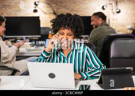 Smiling black woman working on laptop and talking on phone in office Stock Photo