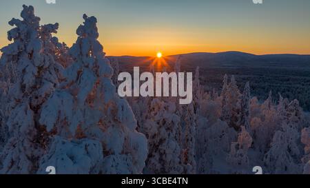 Aerial view of the snow-laden forest with the sun peeking through the horizon, creating a stark contrast between the bright sky and the shadowed trees, Salla, Lapland, Finland. Stock Photo