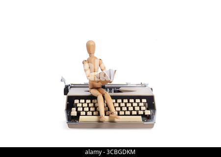A wooden mannequin sitting on a typewriter with a book on his hands Stock Photo
