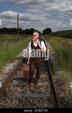 Mid-aged man walks with brown leather suitcase and jacket on railway tracks to the next destination, tall grass and threatening cloud backdrop Stock Photo