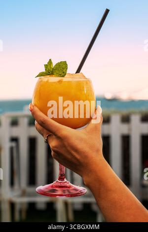 A close-up of a woman's hand holding a large glass of a frozen orange cocktail, garnished with mint, against a beautiful pastel sunset sky at a beach Stock Photo