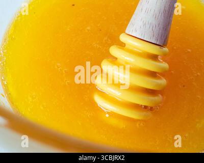Close-up of honey dipper dripping with golden honey in a glass bowl, creating small air bubbles and showcasing the viscous texture of the liquid Stock Photo