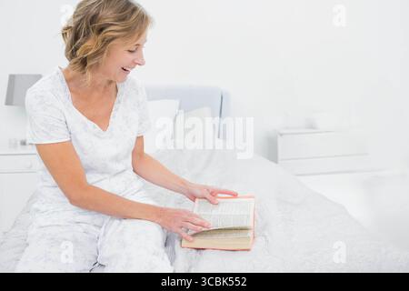Middle-aged woman reading book, turning page on quilted bed by white nightstand with grey lamp. Cozy, relaxation, interior, lifestyle, peaceful, comfo Stock Photo