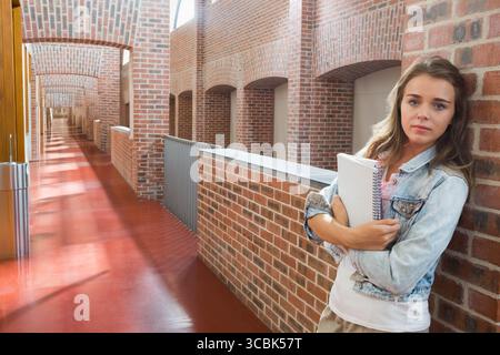 Female student leaning against hallway wall, holding spiral-bound notebooks near railing, trash bin. Educational, academic, hallway, architecture, ref Stock Photo