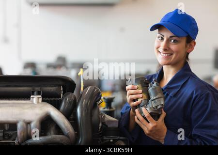 Female mechanic holding starter motor by stripped car engine in garage in blue coverall, copy space. Automotive, repair, industrial, engineering, tool Stock Photo