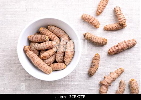 Fresh turmeric rhizomes, in a white bowl, on linen. Underground stems of the turmeric plant Curcuma longa, with bright orange flesh. Stock Photo