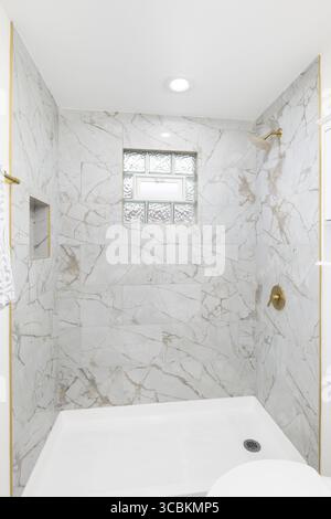 A walk-in shower with marble tiled walls, gold shower head, and glass block window. Stock Photo