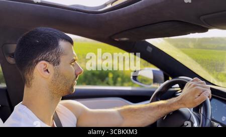 Profile of young handsome man driving a modern electric car with sunlight at background. Young male driver riding on electrical vehicle at countryside Stock Photo