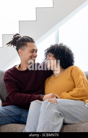 African American couple sitting on grey sofa in living room, gazing at each other near window. Romantic, modern, cozy, lifestyle, interior, affectiona Stock Photo