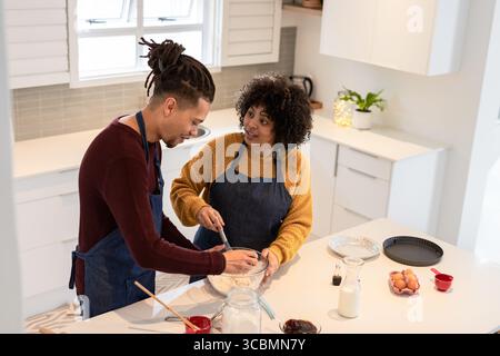 African American couple wearing denim aprons mixing batter at kitchen island with whisk and spatula. Domestic, culinary, contemporary, lifestyle, cozy Stock Photo