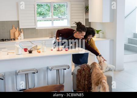 Diverse couple wearing aprons baking at kitchen island with baking tray while petting dog. Home cooking, culinary, domestic, lifestyle, modern design, Stock Photo