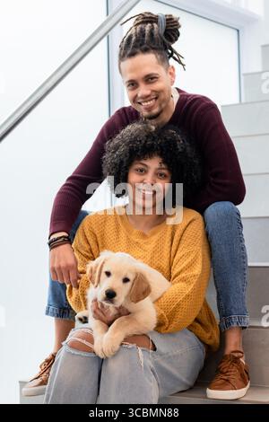 Couple sitting on staircase near frosted window and metal railing holding golden retriever puppy. Companionship, domestic, cozy, contemporary, happine Stock Photo