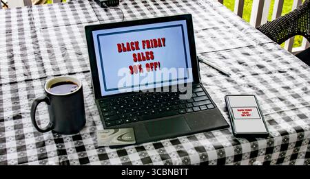 A black checkered table is set up outdoors featuring a laptop displaying a Black Friday sale announcement, a coffee cup, and a smartphone nearby. The Stock Photo