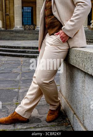 A man in a beige suit and brown vest leans against a stone wall, with his hand in his pocket. He is wearing brown shoes. Stock Photo