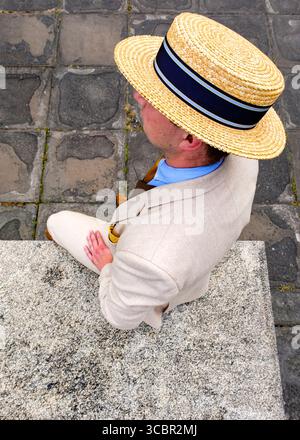 A man wearing a straw hat, light-colored suit, and tie is sitting on a stone step. Stock Photo