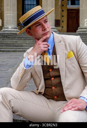 A man in a straw hat and a suit is sitting on the steps of a building, resting his chin on his hand. Stock Photo
