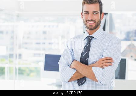 Man in checked shirt and tie crossing arms smiling in office near computer monitors, copy space Stock Photo