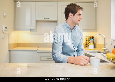 Man wearing button-up shirt, sitting on countertop in kitchen, holding mug near sink, fruit bowl Stock Photo
