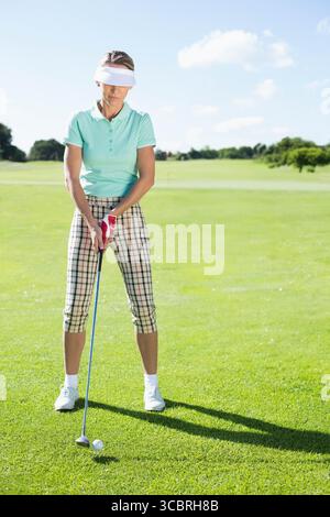 Golf ball sitting beside blue shafted putter on putting green near distant flagstick under sky Stock Photo