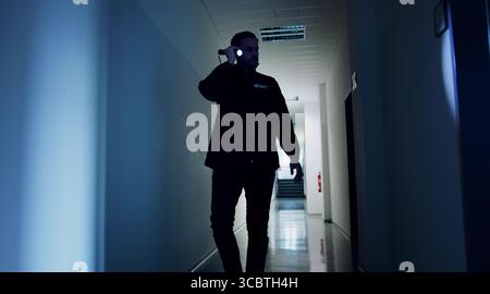 Night Warehouse Security Guard Patrols In Uniform With Flashlight, Ensuring Safety Stock Photo