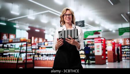Smiling Lady Cashier Assisting Customers In Busy Supermarket Retail Store. Stock Photo