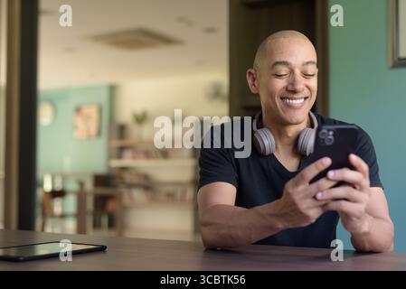 Bald hispanic man in casual black t-shirt seated by window in library, using smartphone. Modern indoor setting with bookshelves, academic atmosphere, Stock Photo