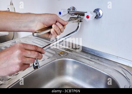 Mounting new sink faucet with straight spout while plumber uses adjustable spanner to secure plumbing connections over clean kitchen sink. Stock Photo