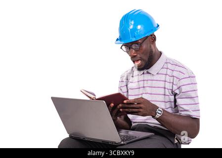 young man engineer wearing hard hat sitting on chair and checking notes in notebook using laptop. Stock Photo
