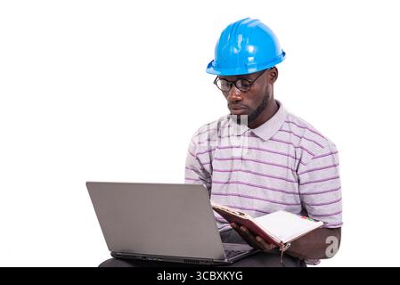young man engineer wearing hard hat sitting on chair and checking notes in notebook using laptop. Stock Photo