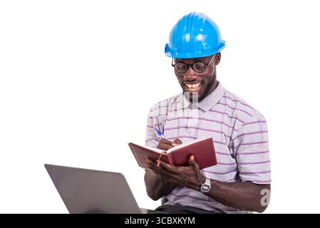 young man engineer wearing hard hat sitting on chair and taking notes in notebook using laptop. Stock Photo