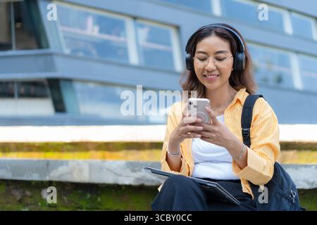 A woman is sitting on a ledge with her phone and laptop. She is wearing headphones and she is focused on her phone Stock Photo