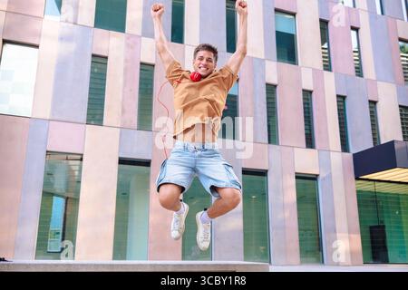 Happy young man jumping outdoors with arms raised in front of modern building Stock Photo