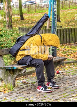 Young man sat on park bench with guitar on back and looking at smartphone - Brussels, Belgium. Stock Photo