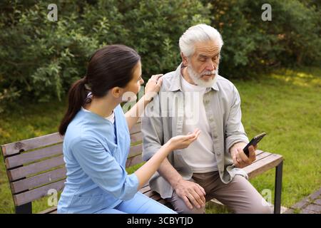 Nurse and elderly man using smartphone on bench outdoors Stock Photo