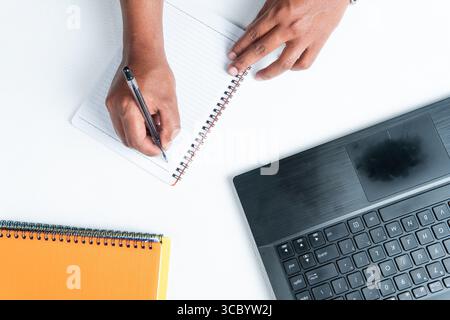 Top view of a man writing in a binder, with an open laptop and a book on a white desk. Perfect for business, study, productivity, and workspace-relate Stock Photo