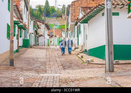 A couple walks hand-in-hand through the picturesque cobblestone streets of Monguí, Boyacá, surrounded by colonial architecture and green accents. Stock Photo