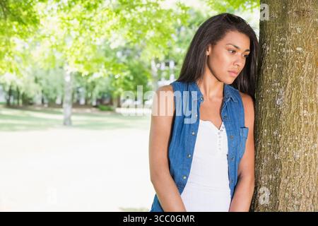 Woman leaning against tree trunk in sunlit park, gazing toward right with sunglasses, copy space Stock Photo