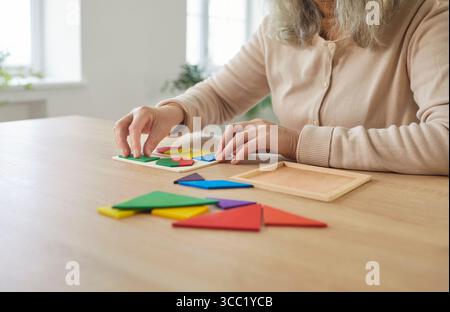 Senior woman sitting playing at table close up, arranging colorful puzzle pieces, working on tangram Stock Photo
