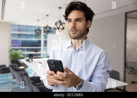 Serious millennial office worker standing in meeting room holding phone Stock Photo