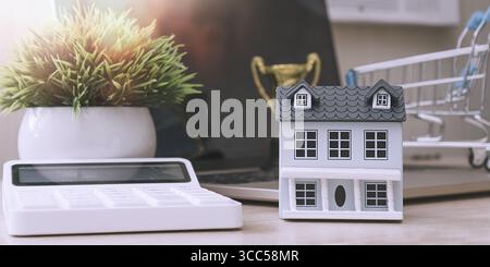 Real estate investment concept featuring a small house model, calculator, shopping cart, and trophy on a bright office desk. Stock Photo