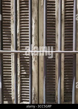 Close-up of an old, weathered window with closed wooden shutters and rustic iron bars, showcasing a textured background of peeling paint. Stock Photo