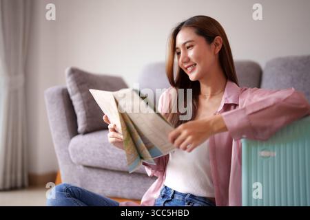 Smiling Asian woman plans vacation using paper map. She prepares for global adventure from cozy home setting. Happy journey inspiration. Stock Photo